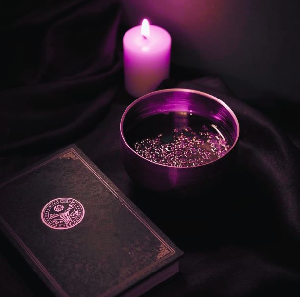 Goldene Klangschale neben einer brennenden Kerze und einem Buch mit Scriptorium-Siegel in pflaumenfarbenem Licht.