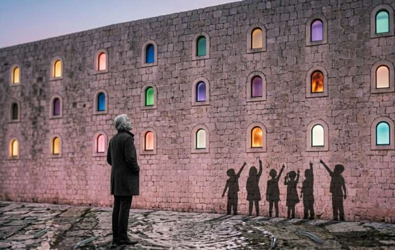 22 Energien Matrix: Ältere schwarz-weiße Frau blickt auf eine Steinmauer mit 22 leuchtenden Fenstern, kindliche Schatten zeigen auf einzelne Fenster