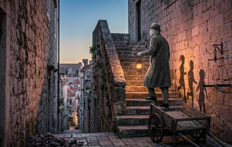 Archetypen auf der Leinwand: Älterer schwarz-weißer Mann steigt mit einer Laterne eine Steintreppe hinauf, unten ein Holzkarren, an der Mauer eine Waage, kindliche Schatten an der rosa-violetten Wand