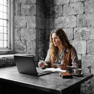 Frau im Boho-Stil sitzt in einem alten Burgzimmer am Eichentisch, hält Kreditkarte und Geldschein in den Händen – Symbolbild für den persönlichen Finanzcode