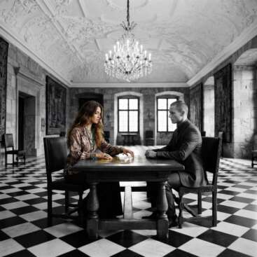Eine Frau im Boho-Stil und ein Mann in Schwarz-Weiß sitzen sich an einem Eichentisch mit goldenen Tarotkarten gegenüber, in einem prunkvollen Burgzimmer mit Schachbrettboden – Symbolbild für den Matrix of Destiny Aufbaukurs