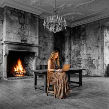 Frau im Boho-Stil sitzt mit Laptop vor einem bunten Kaminfeuer in einem alten Burgzimmer mit Schwarz-Weiß-Steinwänden