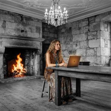 Frau im Boho-Stil hört am Laptop aufmerksam zu, im Hintergrund ein bunt leuchtender Kamin in einem Schwarz-Weiß-Burgzimmer