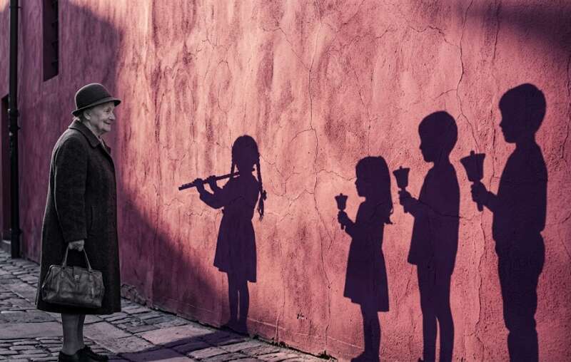 Eine ältere Frau mit Hut steht vor einer rosa-violetten Wand, auf der der Schatten eines Kindes mit Flöte und drei Schatten mit Glocken zu sehen sind